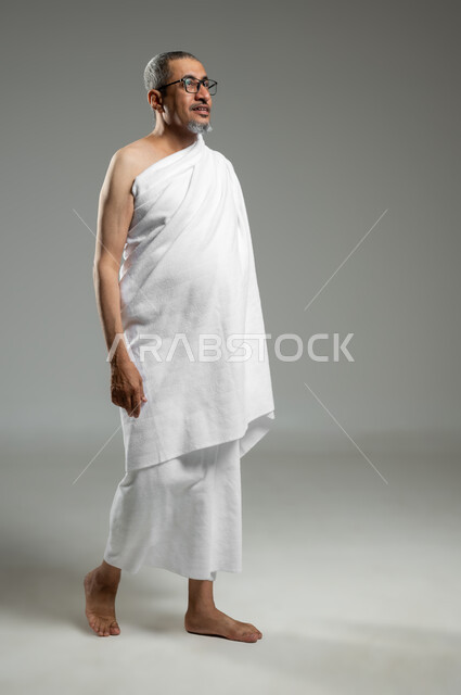 Worshiping and getting closer to God with good deeds, a portrait of a smiling Saudi Gulf Arab man wearing Ihram clothing preparing to perform Hajj and Umrah rituals, full length body, gray background, sensitive use, sensitive content