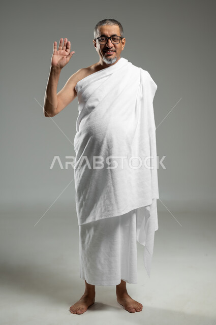 النظر إلى الكاميرا، أداء مناسك الحج والعمرة، بورتريه لرجل عربي سعودي خليجي مبتسم يرتدي ملابس الاحرام ويرفع يده للأعلى لإلقاء التحية والسلام بتعابير السرور، الجسم بالطول الكامل، خلفية رمادية، استخدام حساس, محتوى حساس