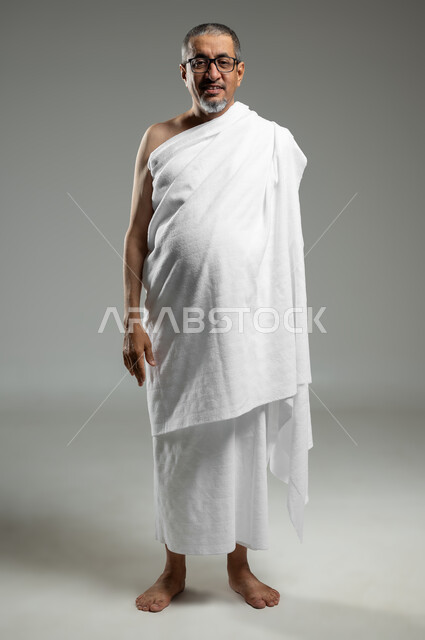 الوقوف باستقامة والنظر الى الكاميرا، الاستعداد لأداء مناسك الحج ، بورتريه لرجل عربي خليجي سعودي مبتسم يرتدي لباس الاحرام يقوم بزيارة بيت الله الحرام في مكة المكرمة، العبادة والتقرب الى الله، الجسم بالطول الكامل، خلفية رمادية، استخدام حساس, محتوى حساس