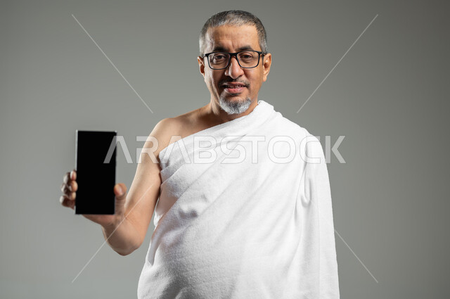 استخدام تطبيقات دليل الحاج والمعتمر على الهاتف المحمول، النظر الى الكاميرا بتعابير السرور ,عرض شاشة سوداء فارغة عبر الجوال، بورتريه مقرب لرجل مسلم خليجي عربي سعودي يرتدي ملابس الاحرام يحمل بيده الهاتف المحمول، خلفية رمادية،  استخدام حساس, محتوى حساس