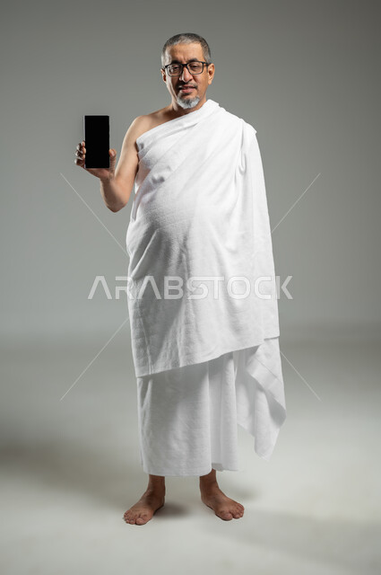 النظر الى الكاميرا بتعابير السرور ,عرض شاشة سوداء فارغة عبر الجوال،  بورتريه لرجل مسلم خليجي عربي سعودي يرتدي ملابس الاحرام يحمل بيده الهاتف المحمول، أداء مناسك الحج والعمرة، استخدام التقنيات الحديثة، الجسم بالطول الكامل، خلفية رمادية، استخدام حساس, محتوى حساس