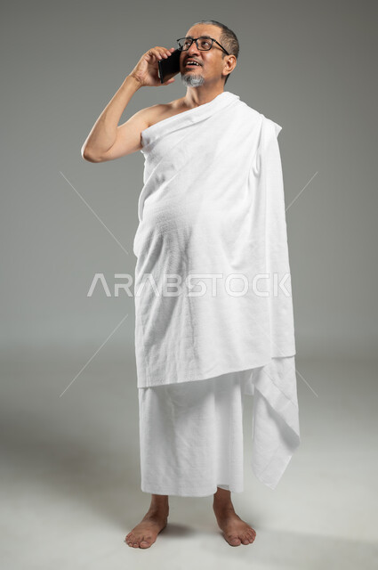 أداء مناسك الحج والعمرة، التواصل مع العائلة والأصدقاء، زيارة بيت الله الحرام في مكة المكرمة، بورتريه مقرب لحاج عربي خليجي سعودي مبتسم يرتدي ملابس الاحرام يقوم بإجراء مكالمة صوتية عبر الهاتف , الجسم بالطول الكامل، خلفية رمادية، استخدام حساس, محتوى حساس