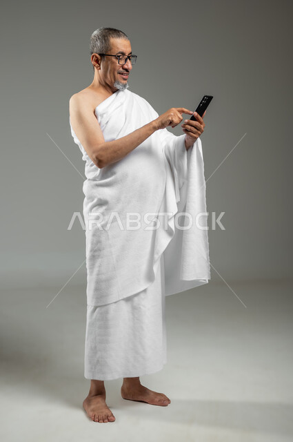 تطبيق مرشد الحج الالكتروني، استخدام خدمة تطبيق الارشادات لأداء مناسك الحج والعمرة، بورتريه لرجل مسلم خليجي عربي سعودي مبتسم يرتدي ملابس الإحرام يستخدم الهاتف المحمول،  الجسم بالطول الكامل، خلفية رمادية، استخدام حساس, محتوى حساس