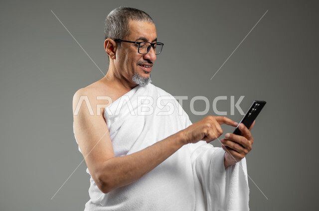 استخدام خدمة تطبيق الارشادات لأداء مناسك الحج والعمرة، بورتريه مقرب لرجل مسلم خليجي عربي سعودي مبتسم يرتدي ملابس الإحرام يستخدم الهاتف المحمول، تطبيق مرشد الحج الالكتروني، خلفية رمادية، استخدام حساس, محتوى حساس