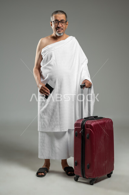 الاستعداد للسفر لأداء مناسك الحج والعمرة، النظر الى الكاميرا بإيماءات السعادة للوصول لمكة المكرمة، بورتريه لرجل مسلم خليجي عربي سعودي مبتسم يرتدي ملابس الاحرام يمسك بيده حقيبة حمراء وجواز السفر، الجسم بالطول الكامل، خلفية رمادية، استخدام حساس, محتوى حساس