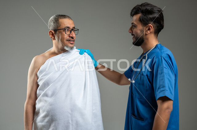 أداء مناسك الحج والعمرة، تقديم الخدمات الصحية لحجاج بيت الله الحرام، الاهتمام بصحة المعتمرين، بورتريه مقرب لممرض اسعافات اولية يرتدي اليونيفورم يضع يده على كتف رجل كبير السن عربي خليجي سعودي يرتدي زي الإحرام، خلفية رمادية،  استخدام حساس, محتوى حساس