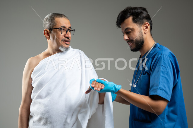 تقديم الخدمات الطبيه لحجاج بيت الله الحرام ، الاهتمام بصحة المعتمرين، بورتريه مقرب لممرض اسعافات اولية يرتدي اليونيفورم يقوم بفحص يد رجل عربي خليجي سعودي يرتدي زي الإحرام، أداء مناسك الحج والعمرة، خلفية رمادية، استخدام حساس, محتوى حساس