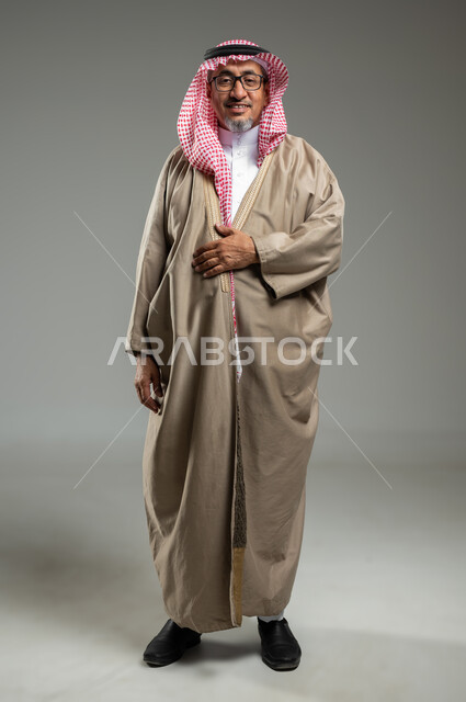 مفهوم الاهتمام في المظهر الخارجي، النظر إلى الكاميرا بإيماءات السرور والفرح، بورتريه لرجل عربي سعودي خليجي مبتسم يرتدي البشت والشماغ التقليدي،  الجسم بالطول الكامل، خلفية رمادية، استخدام حساس، محتوى حساس