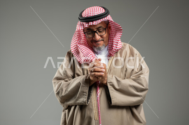 الحمد والشكر وطلب العون , تعابير تدل على احتواء شيء ما بين يديه، الرغبة في تحقيق الأمنيات، بورتريه لرجل عربي خليجي يرتدي الزي السعودي التراثي والشماغ التقليدي، خلفية رمادية، استخدام حساس، محتوى حساس