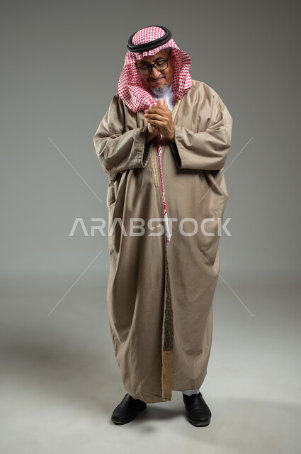 تعابير تدل على احتواء شيء ما بين يديه، الرغبة في تحقيق الأمنيات، بورتريه لرجل عربي خليجي يرتدي الزي السعودي التراثي والشماغ التقليدي ،  الحمد والشكر وطلب العون , الجسم بالطول الكامل، خلفية رمادية، استخدام حساس، محتوى حساس