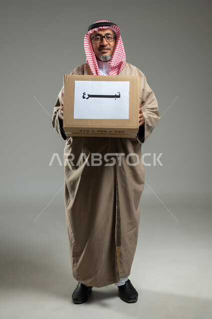 الأعمال الخيرية الإسلاميه في السعودية ،المساعدات والتبرعات الإنسانية، بورتريه مقرب لرجل عربي سعودي خليجي يرتدي الزي التراثي التقليدي يمسك بيده صندوق كرتوني، النظر إلى الكاميرا بإيماءات السرور، الجسم بالطول الكامل، خلفية رمادية، استخدام حساس، محتوى حساس
