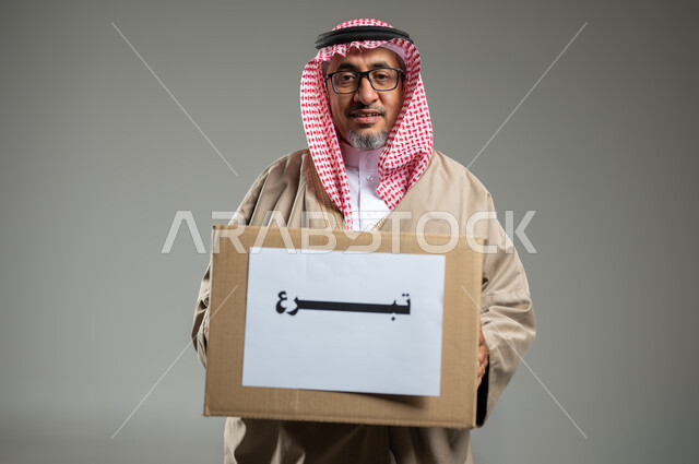 المساعدات والتبرعات الإنسانية، مفهوم التبرع والأعمال الخيرية الإسلاميه في السعودية ، بورتريه مقرب لرجل عربي سعودي خليجي يرتدي الزي التراثي التقليدي يمسك بيده صندوق كرتوني، النظر إلى الكاميرا بإيماءات السرور، خلفية رمادية، استخدام حساس، محتوى حساس