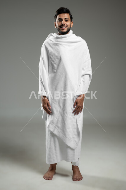 الوقوف باستقامة والنظر إلى شيء معين، النظر إلى الكاميرا بتعابير السرور، بورتريه لرجل مسلم عربي سعودي خليجي يرتدي ملابس الإحرام يقوم بزيارة بيت الله الحرام في مكة المكرمة، الجسم بالطول الكامل، خلفية رمادية، استخدام حساس، محتوى حساس