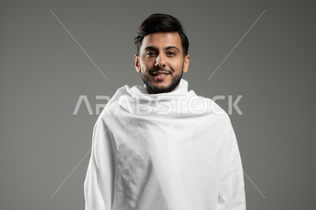 النظر إلى الكاميرا بإيماءات السرور، بورتريه مقرب لشاب عربي سعودي خليجي مبتسم يرتدي ملابس الإحرام يقوم بزيارة بيت الله الحرام في مكة المكرمة، خلفية رمادية،  استخدام حساس، محتوى حساس