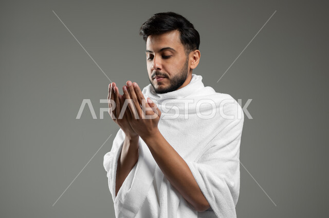 أداء العمرة في مكة المكرمة ، التقرب الى الله بالصلاة والعبادة، بورتريه مقرب لشاب مسلم عربي سعودي خليجي يرتدي ملابس الاحرام يرفع يديه للأعلى بإيماءات الدعاء، خلفية رمادية، استخدام حساس، محتوى حساس