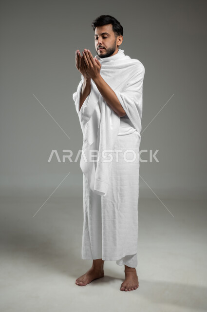 Getting closer to God through prayer and worship, a portrait of a Saudi Gulf Arab young man wearing Ihram clothes, raising his hands up and calling upon God, performing Hajj and Umrah rituals, full length body, gray background, sensitive use, sensitive content