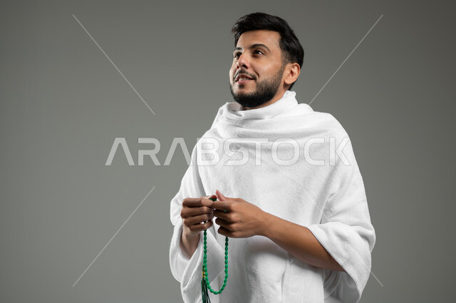 أداء العمرة في مكة المكرمة، مفهوم العبادة والتقرب الى الله، تسبيح وأذكار وأدعية، بورتريه مقرب لرجل مسلم خليجي عربي سعودي مبتسم يرتدي ملابس الإحرام ويحمل سبحة خضراء، خلفية رمادية،  استخدام حساس، محتوى حساس