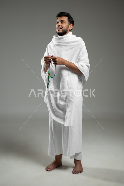 The concept of worship and drawing closer to God, praises, remembrances and supplications, performing Umrah in Mecca, a portrait of a smiling Gulf Arab Saudi Muslim man wearing Ihram clothes and carrying a green rosary, full-length body, gray background, sensitive use, sensitive content