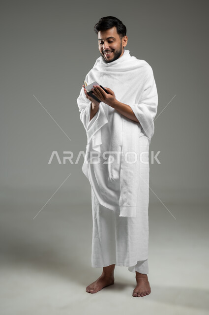 Expressions of enthusiasm and happiness for performing Umrah rituals, preparing to go for Hajj, portrait of a young Gulf Arab Saudi Muslim wearing Ihram clothing and holding a passport in his hand, full-length body, gray background, sensitive use, sensitive content