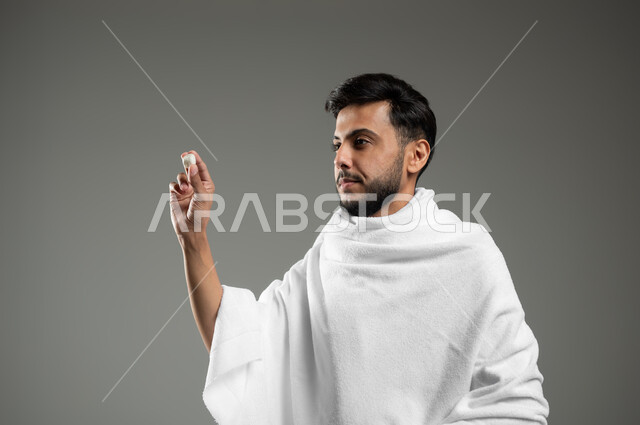 العبادة والتقرب الى الله، رمي جمرة العقبة في يوم النحر، بورتريه مقرب لشاب عربي سعودي خليجي يرتدي ملابس الاحرام يحمل بيده حصوى للقيام برجم ابليس، أداء مناسك الحج والعمرة، خلفية رمادية، استخدام حساس، محتوى حساس