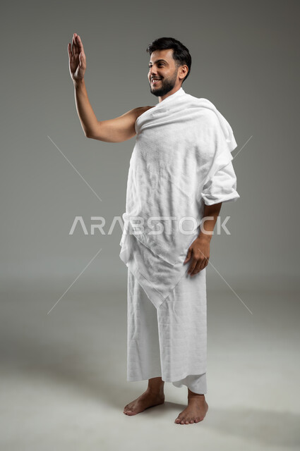 أداء مناسك الحج والعمرة، بورتريه لشاب عربي سعودي خليجي مبتسم يرتدي ملابس الاحرام ويرفع يده للأعلى لإلقاء التحية والسلام بتعابير السرور، العبادة والتقرب الى الله، الجسم بالطول الكامل، خلفية رمادية، استخدام حساس، محتوى حساس