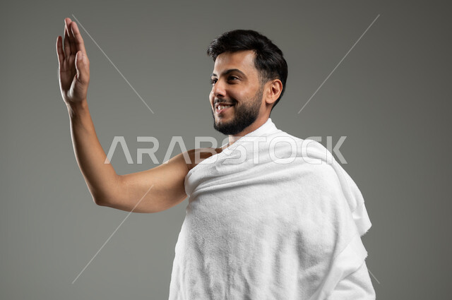العبادة والتقرب الى الله، أداء مناسك الحج والعمرة، بورتريه مقرب لشاب عربي سعودي خليجي مبتسم يرتدي ملابس الاحرام ويرفع يده للأعلى لإلقاء التحية والسلام بتعابير السرور، خلفية رمادية،  استخدام حساس، محتوى حساس