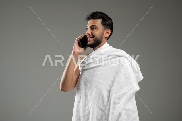 Communicating with family and friends, visiting the Holy House of God in Mecca, a close-up portrait of a smiling Saudi Gulf Arab young man wearing Ihram clothing making a voice call via mobile phone, performing Hajj and Umrah rituals, gray background, sensitive use, sensitive content