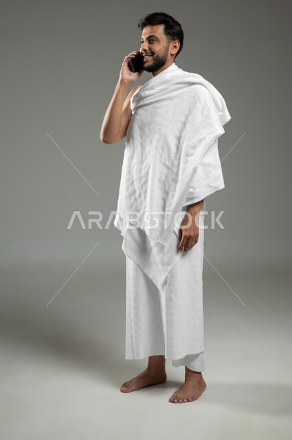 Visiting the Holy House of God in Mecca, communicating with family and friends, portrait of a smiling Saudi Gulf Arab man wearing Ihram clothes making a voice call via mobile phone, performing Hajj and Umrah rituals, full length body, gray background, sensitive use, sensitive content