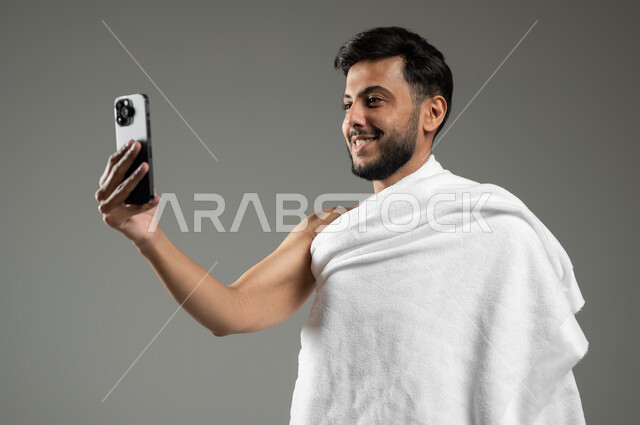 بث مباشر عبر وسائل التواصل الاجتماعي، استخدام أجهزة تقنية حديثة، أداء مناسك الحج والعمرة، بورتريه مقرب لرجل عربي خليجي سعودي مبتسم يرتدي ملابس الاحرام يقوم بالتقاط صورة سيلفي تذكارية من خلال الهاتف المحمول، خلفية رمادية،  استخدام حساس، محتوى حساس