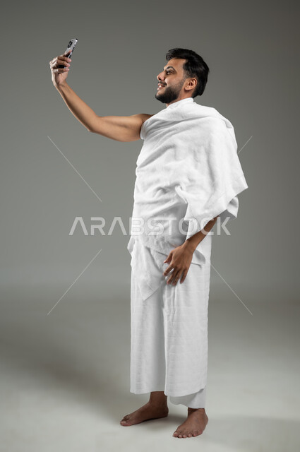 توثيق اللحظات السعيدة في مكة المكرمة، استخدام أجهزة تقنية حديثة، أداء مناسك الحج والعمرة، بورتريه لرجل عربي خليجي سعودي يرتدي ملابس الاحرام ويقوم بالتقاط صور سيلفي تذكارية عبر الهاتف المحمول، الجسم بالطول الكامل، خلفية رمادية، استخدام حساس، محتوى حساس