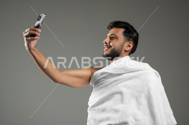 استخدام أجهزة تقنية حديثة، أداء مناسك الحج والعمرة، بورتريه مقرب لرجل عربي خليجي سعودي يرتدي ملابس الاحرام يقوم بالتقاط صور سيلفي تذكارية عبر الهاتف المحمول، توثيق اللحظات السعيدة في مكة المكرمة، خلفية رمادية، استخدام حساس، محتوى حساس