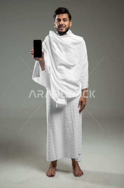 عرض شاشة سوداء فارغة عبر الجوال، النظر الى الكاميرا بتعابير السرور, عرض تطبيقات دليل الحاج والمعتمر على الهاتف المحمول، بورتريه لشاب مسلم خليجي عربي سعودي يرتدي ملابس الاحرام يحمل بيده الهاتف المحمول، الجسم بالطول الكامل، خلفية رمادية، استخدام حساس، محتوى حساس