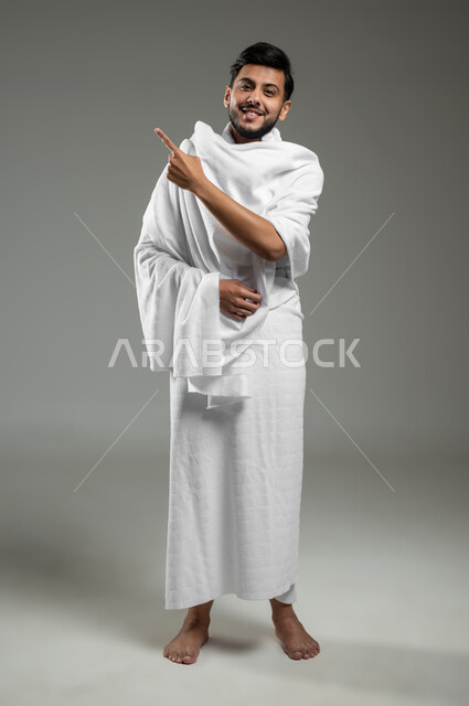 النطر إلى الكاميرا، الوقوف باستقامة و النظر إلى مكان معين، ايماءات السعادة لرؤية شيء ما، بورتريه لشاب مسلم عربي سعودي خليجي يرتدي ملابس الاحرام يشير بأصبع السبابة نحو الأعلى، آداء مناسك الحج والعمرة،  الجسم بالطول الكامل، خلفية رمادية، استخدام حساس، محتوى حساس
