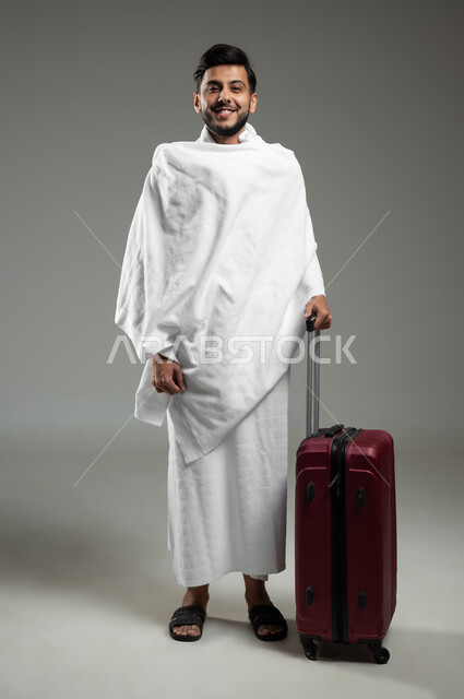 النظر الى الكاميرا بإيماءات السعادة والحماس  للوصول لمكة المكرمة، التأهب والاستعداد للسفر لأداء مناسك الحج والعمرة، بورتريه لرجل مسلم خليجي عربي سعودي مبتسم يرتدي ملابس الاحرام يمسك بيده حقيبة حمراء، الجسم بالطول الكامل، خلفية رمادية، استخدام حساس، محتوى حساس