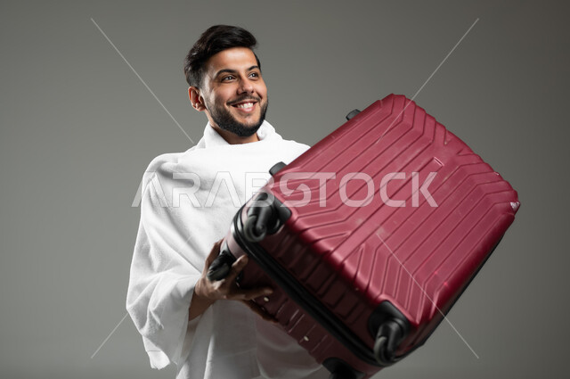 الاستعداد للتوجه و الانطلاق لزيارة بيت الله الحرام, بورتريه مقرب لشاب عربي خليجي سعودي يرتدي ملابس الاحرام و يحمل حقيبة سفر حمراء, أداء فريضة الحج و العمرة , ايماءات السعادة والحماس للوصول لمكة المكرمة, خلفية رمادية، استخدام حساس، محتوى حساس