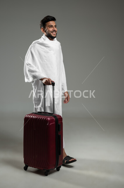 المشي والذهاب لأداء فريضة الحج والعمرة، بورتريه لشاب مسلم خليجي عربي سعودي مبتسم يرتدي ملابس الاحرام ويمسك بيده حقيبة السفر ، ايماءات السعادة والحماس للوصول لمكة المكرمة،   الجسم بالطول الكامل، خلفية رمادية، استخدام حساس، محتوى حساس