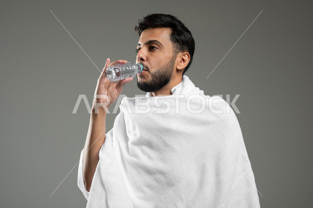 Drink plenty of fluids to keep the body from dehydration, performing Hajj and Umrah rituals, a close-up portrait of a Saudi Gulf Arab man wearing the Ihram dress, drinking fresh water, quenching himself with Zamzam water, gray background, sensitive use, sensitive content