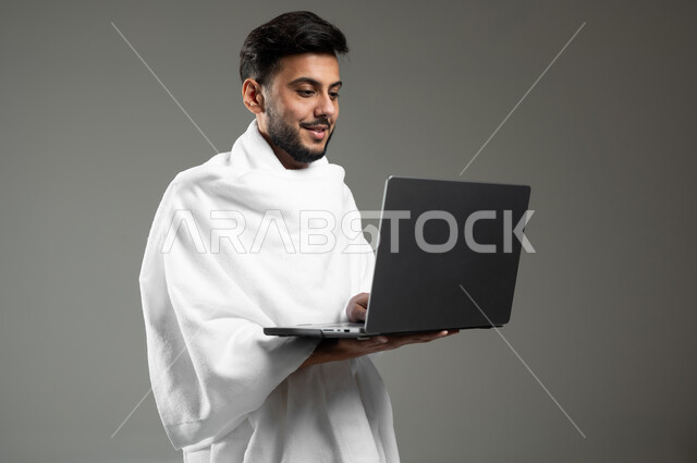 استخدام الأجهزة التقنية الحديثة في تصفح مواقع الارشادات لأداء مناسك الحج والعمرة، بورتريه مقرب لرجل مسلم خليجي عربي سعودي مبتسم يرتدي ملابس الاحرام يحمل بيده جهاز الحاسوب المحمول بإيماءات السرور ، خلفية رمادية، استخدام حساس، محتوى حساس
