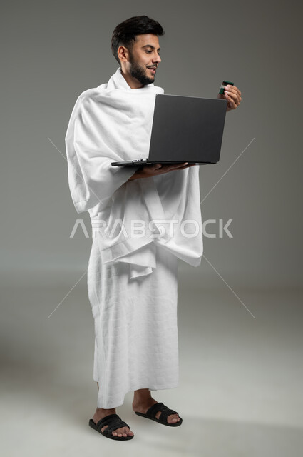 الحجز الالكتروني في حملات الحج و العمرة, الشراء و الدفع عن طريق بطاقة الصراف الإئتمانية, بورتريه لرجل خليجي سعودي عربي يرتدي ملابس الاحرام يحمل بيده الحاسوب المحمول وكرت البنك المصرفي,  الجسم بالطول الكامل، خلفية رمادية، استخدام حساس، محتوى حساس