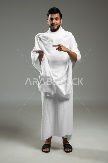 النظر إلى الكاميرا بإيماءات السرور ،بورتريه لرجل عربي خليجي سعودي مبتسم يرتدي ملابس الإحرام يرفع يده للأعلى لعرض شيء ما، الإشارة بإصبع السبابة إلى شيء معين، أداء مناسك الحج والعمره ، الجسم بالطول الكامل، خلفية رمادية، استخدام حساس، محتوى حساس