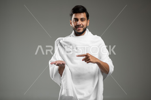 الإشارة بإصبع السبابة إلى شيء معين، أداء مناسك الحج والعمره ،النظر إلى الكاميرا بإيماءات السرور ،بورتريه لرجل عربي خليجي سعودي مبتسم يرتدي ملابس الإحرام يرفع يده للأعلى لعرض شيء ما، خلفية رمادية، استخدام حساس، محتوى حساس