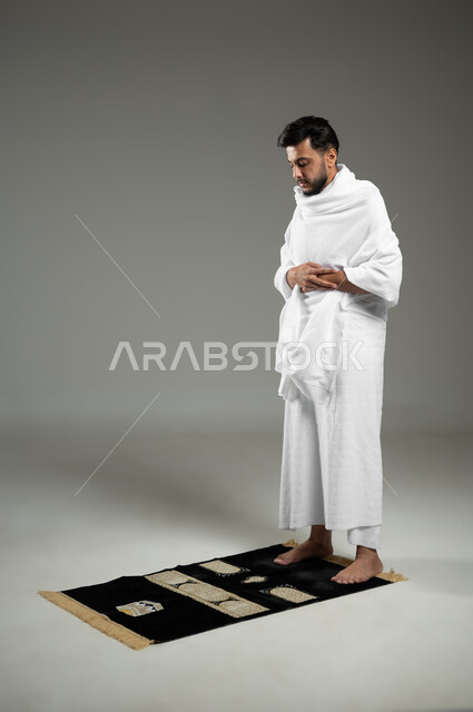 Praying and supplicating to God, performing the rituals of Hajj and Umrah, worship and drawing closer to God, a portrait of a Saudi Arabian Gulf pilgrim wearing Ihram clothing and performing the obligatory prayer, kneeling and prostrating, remembrances and supplications, full-length body, gray background, sensitive use, sensitive content