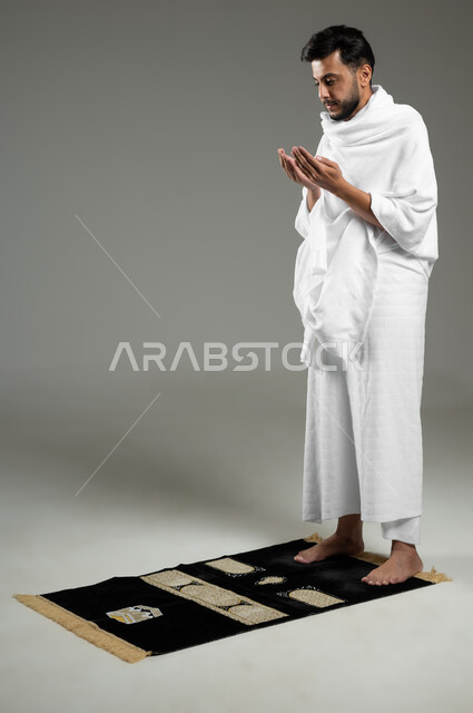 الدعاء والعباده والتقرب الى الله، بورتريه لحاج عربي سعودي خليجي يرتدي ملابس الاحرام يرفع يديه لاعلى، آداء مناسك الحج والعمرة، الركن الخامس من أركان الاسلام، الجسم بالطول الكامل، خلفية رمادية،  استخدام حساس، محتوى حساس
