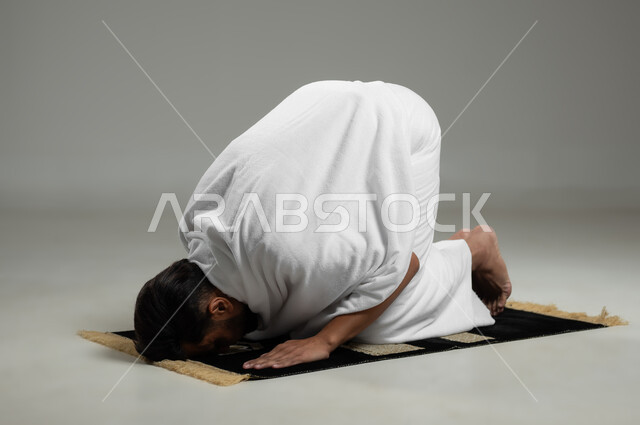 مفهوم العبادة والتقرب إلى الله تعالى ،أداء الفرائض الدينية و مناسك الحج والعمره ،بورتريه لحاج عربي خليجي سعودي يرتدي ملابس الإحرام يقوم بالسجود أثناء الصلاة، خلفية رمادية، استخدام حساس، محتوى حساس