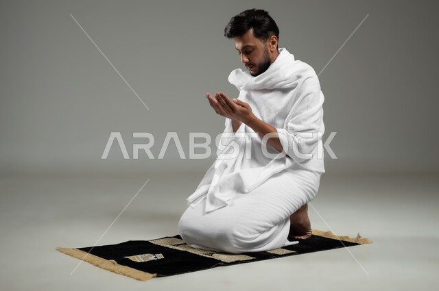 أداء مناسك الحج والعمرة, بورتريه لرجل عربي سعودي خليجي يرتدي لباس الإحرام يجلس على سجادة الصلاة ويرفع يديه إلى الأعلى بإيماءات الدعاء ,  الإكثار من ذكر الله تعالى والإجتهاد في العباده, خلفية رمادية، استخدام حساس، محتوى حساس