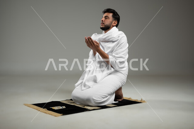الإكثار من ذكر الله تعالى والإجتهاد في العباده, بورتريه لحاج عربي سعودي خليجي يرتدي لباس الإحرام يجلس على سجادة الصلاة ويرفع يديه إلى الأعلى بإيماءات الدعاء , أداء مناسك الحج والعمرة, خلفية رمادية، استخدام حساس، محتوى حساس