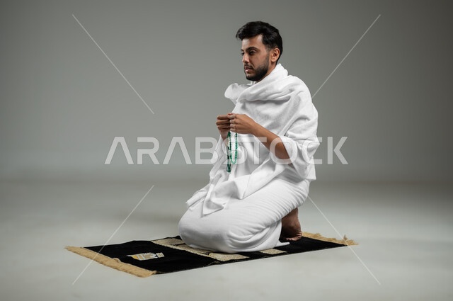 الإجتهاد في العباده والتقرب الى الله، قراءة الأذكار والأدعية، بورتريه لحاج عربي سعودي خليجي يرتدي لباس الإحرام يجلس على سجادة الصلاة يمسك بيده السبحة يقوم بالتسبيح والاستغفار، خلفية رمادية، استخدام حساس، محتوى حساس