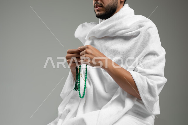 قراءة الأذكار والأدعية، التسبيح والتهليل وذكر الله تعالى، بورتريه مقرب لحاج عربي سعودي خليجي يرتدي لباس الإحرام يمسك بيده السبحة، العبادة والتقرب لرب العالمين، خلفية رمادية، استخدام حساس، محتوى حساس