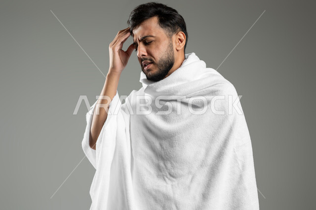 الاستعداد لأداء مناسك الحج والعمرة، الإحساس بألم الرأس والصداع، آلام الشقيقة والصداع النصفي، بورتريه مقرب لحاج عربي خليجي سعودي يرتدي ثياب الإحرام يضع يده على رأسه بايماءات التعب والإرهاق، خلفية رمادية، استخدام حساس، محتوى حساس