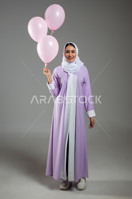 الوقوف باستقامة وثقة والنظر إلى الكاميرا, مفهوم الأناقة والاهتمام بالمظهر الخارجي, بورتريه لشابة عربية خليجية سعودية مبتسمة ترتدي العباءة الملونة والحجاب وتمسك بيدها مجموعة من البلالين الوردية, صورة الجسم بالطول الكامل, خلفية رمادية, محتوى حساس, استخدام حساس